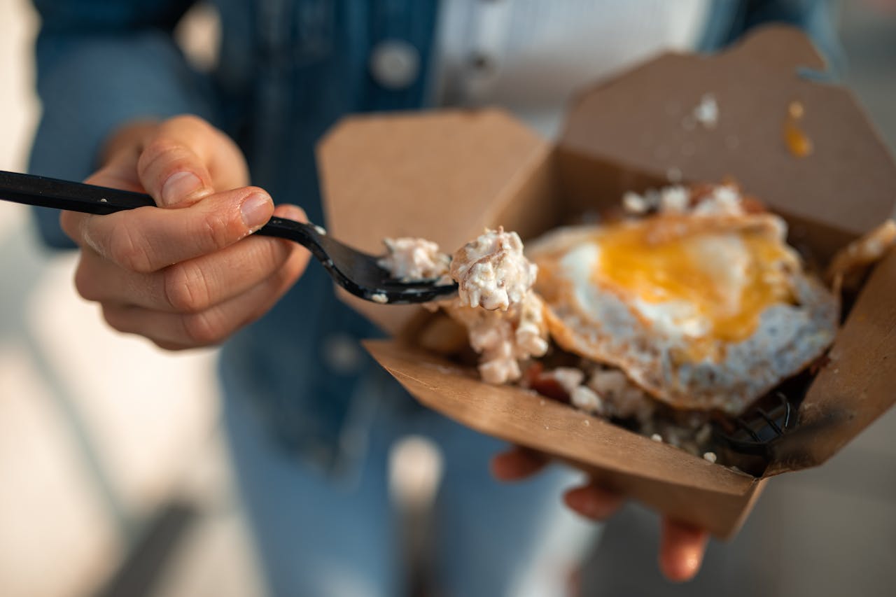 Close-up of a sunny side up egg in a takeout box being eaten with a fork.